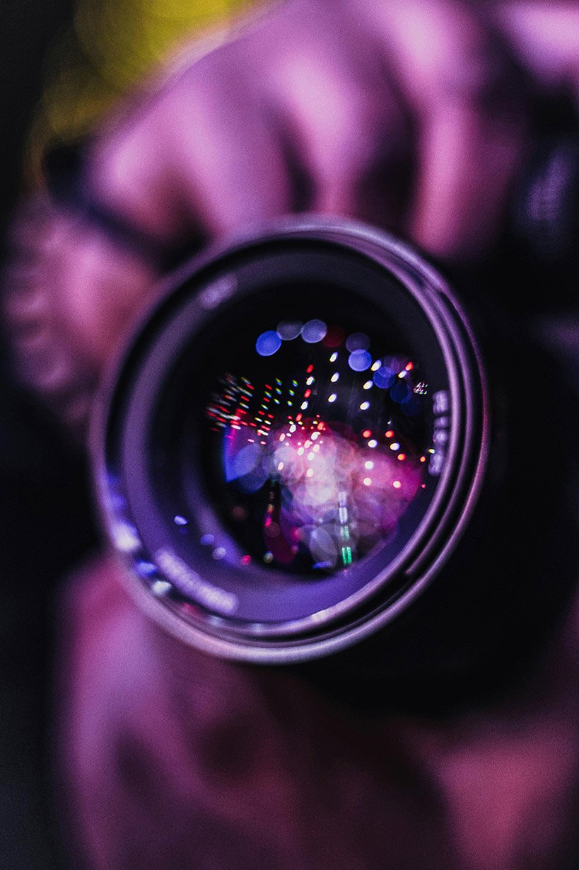 Close-up of a camera lens held to take a shot, with the street lights reflecting in it.